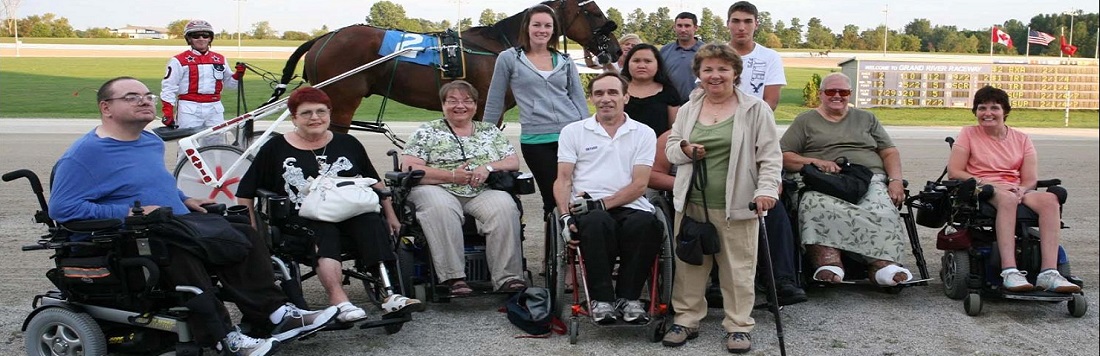 Group of people, some in wheelchairs, posing for a picture in front of the Grand River Raceway with a horse and jockey.
